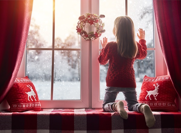 © konstantin yuganov – stock.adobe.com Mädchen im roten Pullover kniet auf Fensterbank und schaut nach dem Rentierschlitten, Weihnachtskranz am Fenster