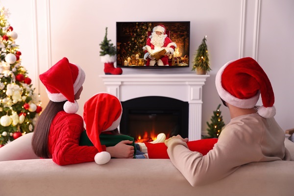 © New Africa / Weihnachtsmann Claudius - stock.adobe.com - Videobotschaft auf dem Fernseher im Wohnzimmer Familie mit Weihnachtsmützen schaut auf einem Fernseher über dem Kamin eine Videobotschaft von Weihnachtsmann Claudius